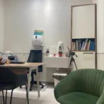 משרד קטן עם שולחן, כיסאות, מדף ספרים, מתקן חיטוי המותקן על הקיר, חצובה וציוד משרדי מגוון.