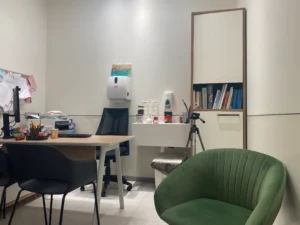 משרד קטן עם שולחן, כיסאות, מדף ספרים, מתקן חיטוי המותקן על הקיר, חצובה וציוד משרדי מגוון.