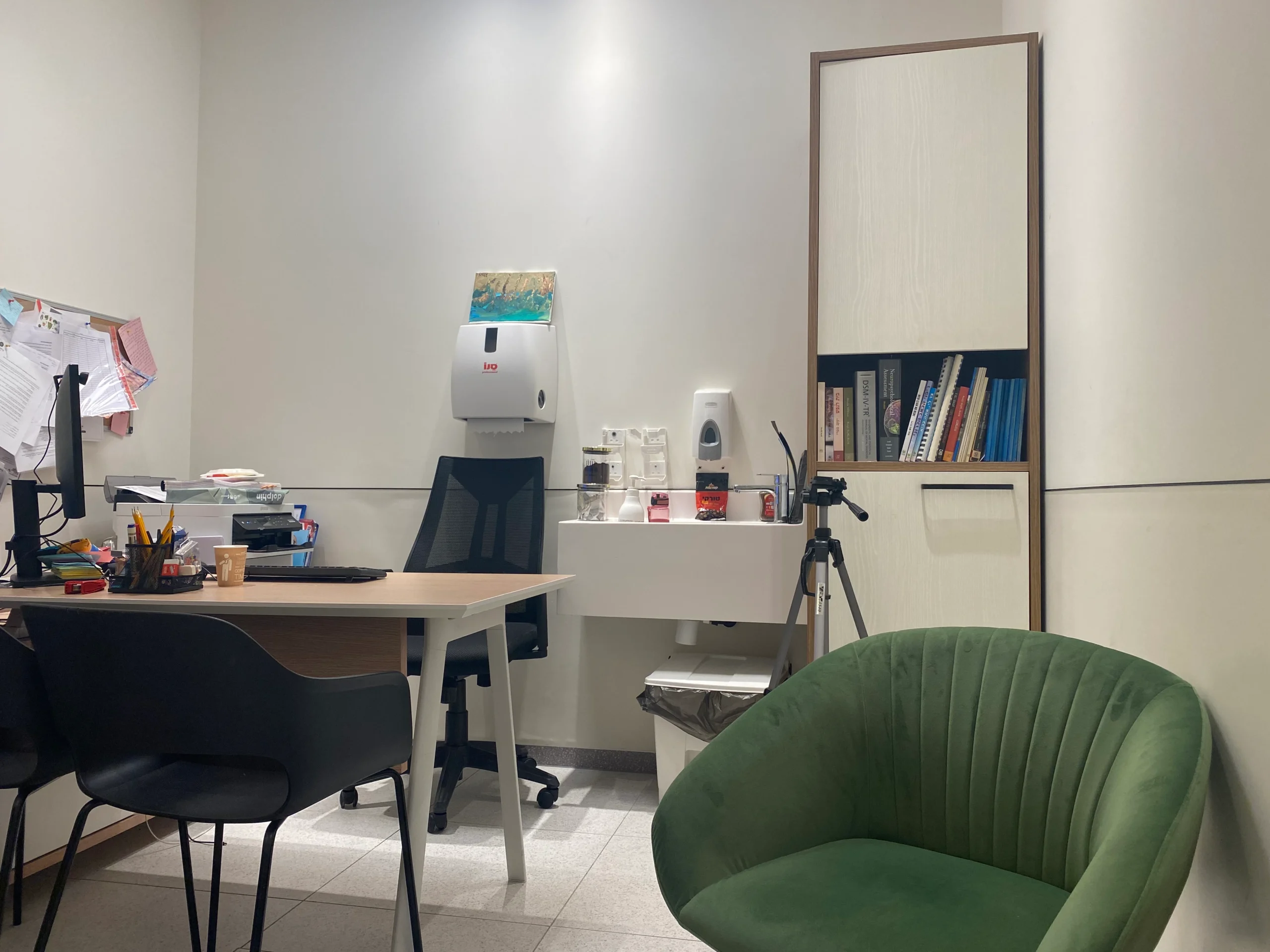 משרד קטן עם שולחן, כיסאות, מדף ספרים, מתקן חיטוי המותקן על הקיר, חצובה וציוד משרדי מגוון.