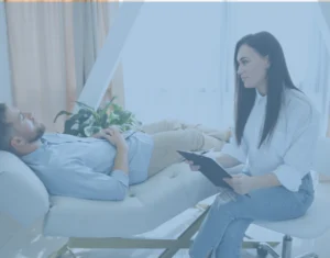 גבר שוכב על ספה בזמן שאישה מחזיקה בלוח כתיבה יושבת לידו, ככל הנראה במהלך טיפול או פגישת ייעוץ.