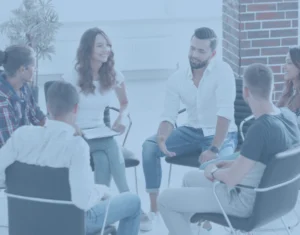 שישה אנשים יושבים במעגל ומשוחחים בחדר מואר ומודרני עם עמוד לבנים וחלונות גדולים.