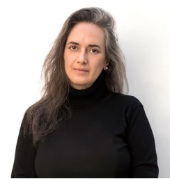 אדם עם שיער אפור ארוך וגלי, לבוש בסוודר שחור עם צווארון גולף, עומד על רקע לבן חלק.