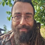 גבר עם משקפיים, זקן מלא וראסטות ארוכות מחייך בחוץ מול עץ עלים ביום שטוף שמש.