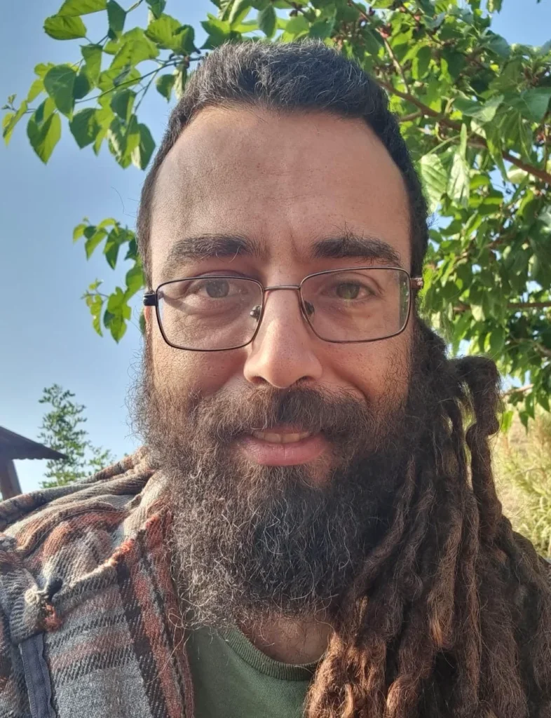 גבר עם משקפיים, זקן מלא וראסטות ארוכות מחייך בחוץ מול עץ עלים ביום שטוף שמש.