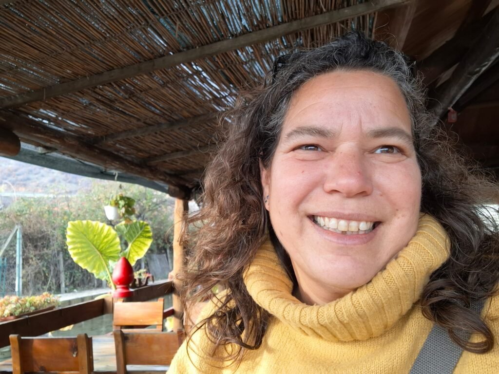 אישה בסוודר צהוב עם צווארון גולף מחייכת למצלמה כשהיא יושבת בחוץ תחת פרגולה מעץ, עם צמחים וכסאות ריקים ברקע.