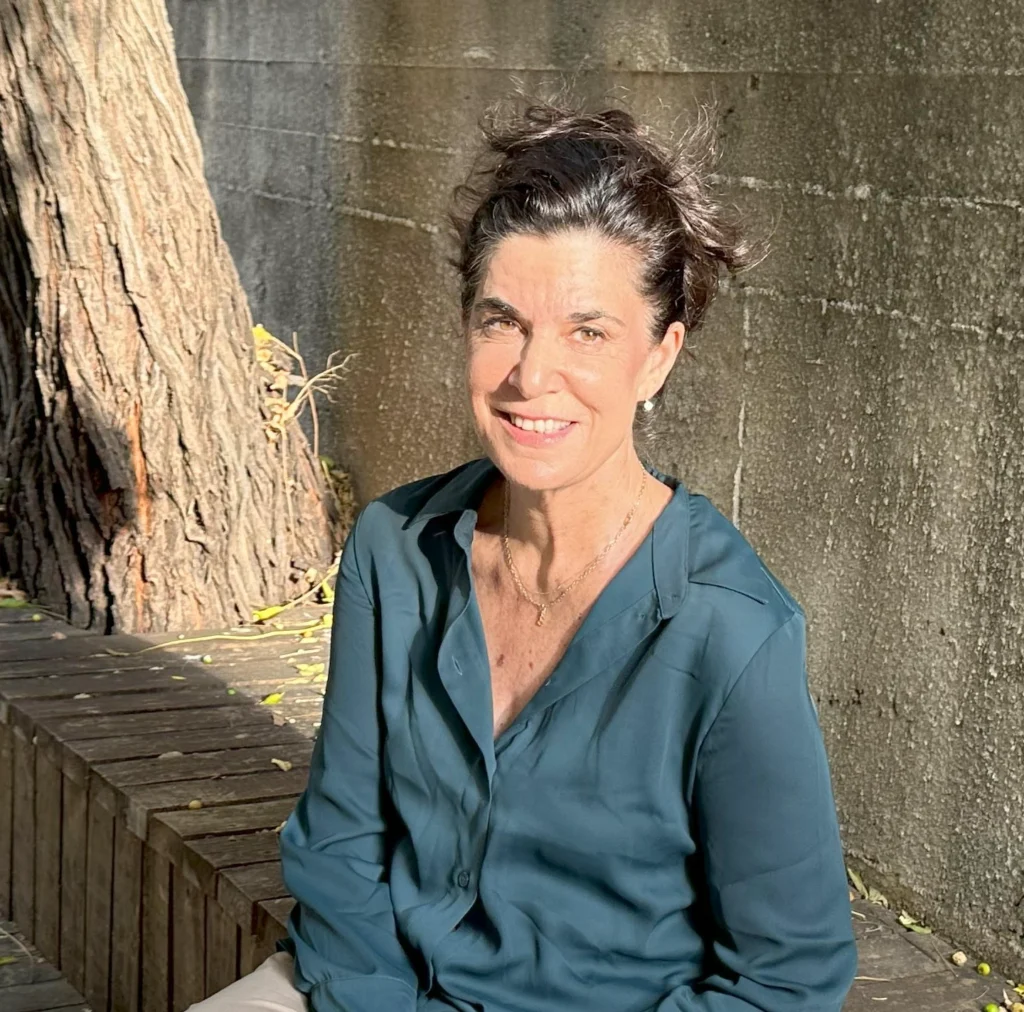 אישה עם שיער קצר וכהה, לבושה בחולצה בצבע טורקיז, יושבת על ספסל עץ בחוץ, מחייכת, כשקרני השמש וצללי העצים מוטלים על קיר הבטון מאחוריה.