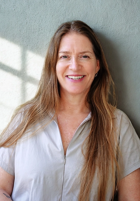 אישה עם שיער חום ארוך, לבושה בחולצה קצרה עם רוכסן בצבע אפור בהיר, מחייכת מול קיר מרקם עם אור שמש וצללים.