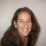 אישה עם שיער חום ארוך, לבושה בחולצה שחורה ועונדת עגילי חישוק, מחייכת למצלמה על רקע בהיר וחלקה.