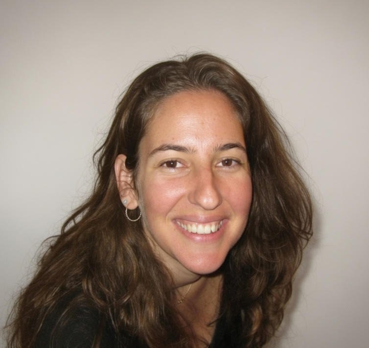 אישה עם שיער חום ארוך, לבושה בחולצה שחורה ועונדת עגילי חישוק, מחייכת למצלמה על רקע בהיר וחלקה.