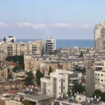 נוף עירוני הכולל בנייני מגורים בינוניים עם גגות שטוחים, עצים הפזורים בין המבנים, והים הנראה ברקע תחת שמיים בהירים.