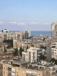 נוף עירוני הכולל בנייני מגורים בינוניים עם גגות שטוחים, עצים הפזורים בין המבנים, והים הנראה ברקע תחת שמיים בהירים.