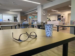 זוג משקפיים וכוס נייר על שולחן עץ בחדר המתנה, עם אנשים יושבים ועובד ברקע.