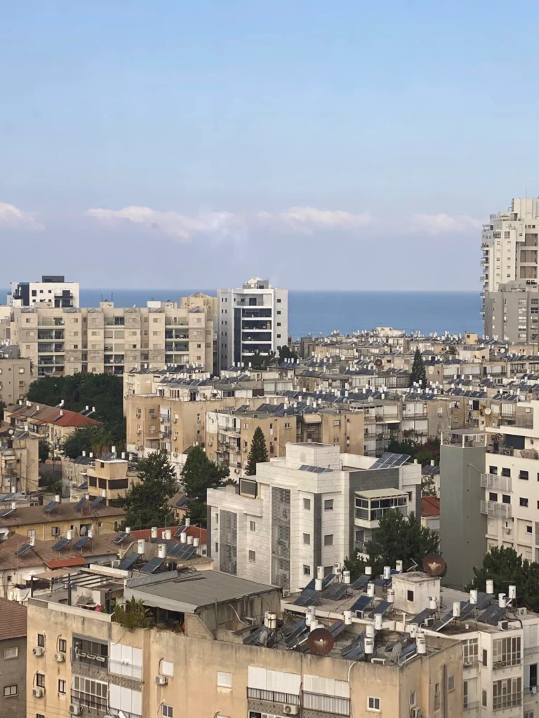 נוף עירוני הכולל בנייני מגורים בינוניים עם גגות שטוחים, עצים הפזורים בין המבנים, והים הנראה ברקע תחת שמיים בהירים.