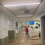 מסדרון בית חולים עם ריצוף בדוגמת עץ, דלפק קבלה לבן מימין, מסכים המותקנים על הקיר, ציוד רפואי ותאורה פלורוסנטית תקרתית.