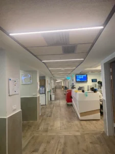 מסדרון בית חולים עם ריצוף בדוגמת עץ, דלפק קבלה לבן מימין, מסכים המותקנים על הקיר, ציוד רפואי ותאורה פלורוסנטית תקרתית.