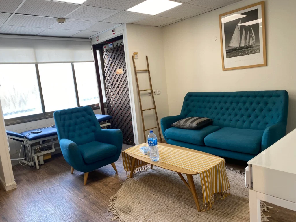 אזור המתנה עם ספה וכורסה בצבע טורקיז, שולחן קפה עם בקבוק מים, מפה מפוספסת, שולחן בדיקה רפואי ותמונה ממוסגרת של סירת מפרש על הקיר.