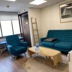 אזור המתנה עם ספה וכורסה בצבע טורקיז, שולחן קפה עם בקבוק מים, מפה מפוספסת, שולחן בדיקה רפואי ותמונה ממוסגרת של סירת מפרש על הקיר.