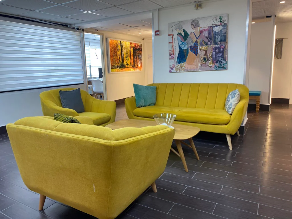 אזור ישיבה מודרני עם ספות צהובות, שולחן קפה עגול מעץ, תמונות קיר וכריות, על רצפת אריחים כהה עם חלונות גדולים וקירות לבנים.