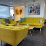 אזור ישיבה מודרני עם ספות צהובות, שולחן קפה עגול מעץ, תמונות קיר וכריות, על רצפת אריחים כהה עם חלונות גדולים וקירות לבנים.