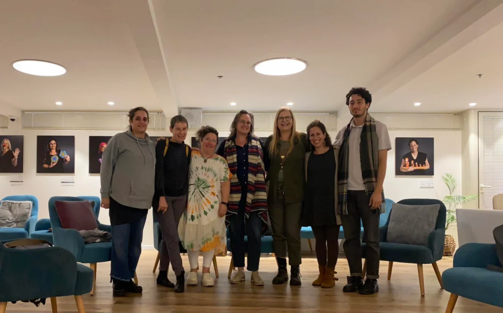 שבעה אנשים עומדים זה לצד זה בחדר מודרני ומואר היטב, עם כיסאות כחולים ותמונות דיוקן המוצגות על הקיר מאחוריהם.