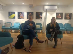 שתי נשים יושבות ומשוחחות בחדר מודרני עם כיסאות כחולים ורצפת עץ, כשמאחוריהן תלויות תמונות דיוקן על הקיר.