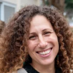 אישה עם שיער חום מתולתל מחייכת למצלמה בחוץ. היא עונדת עגילים צבעוניים ולובשת חולצה כהה.