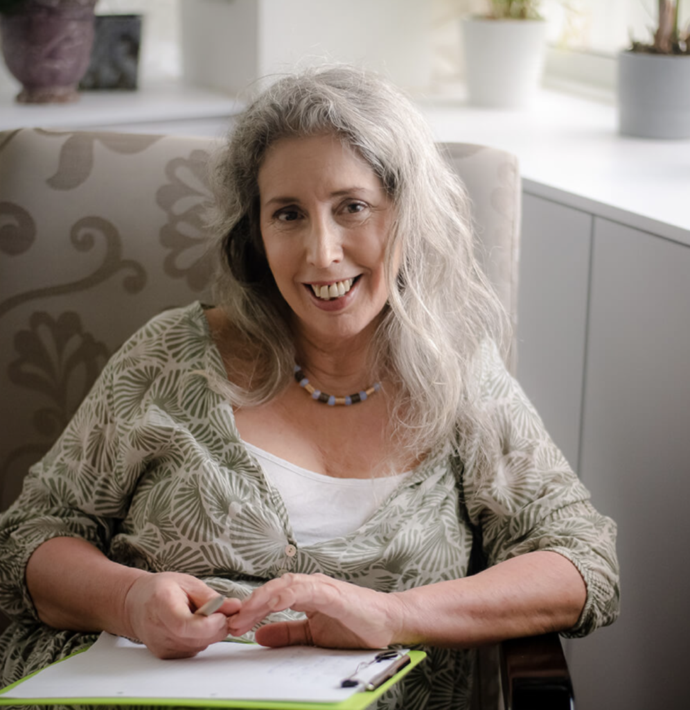 אישה עם שיער אפור יושבת בכורסה, מחייכת למצלמה, אוחזת בעט ובקלסר עם ניירות. תפאורה פנימית עם צמחים ברקע.