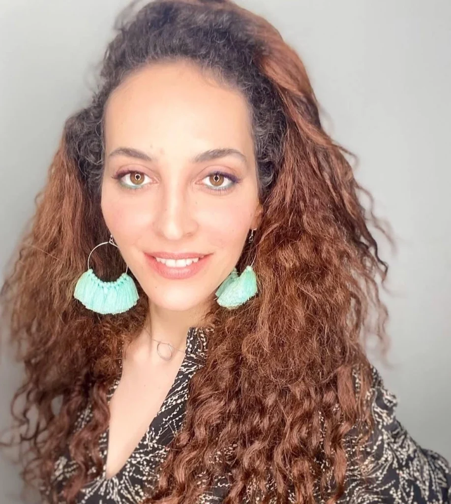 אישה עם שיער חום ארוך ומתולתל, עונדת עגילי ציצית גדולים בצבע תכלת ולובשת חולצה שחורה עם הדפס, מחייכת על רקע בהיר וחלק.