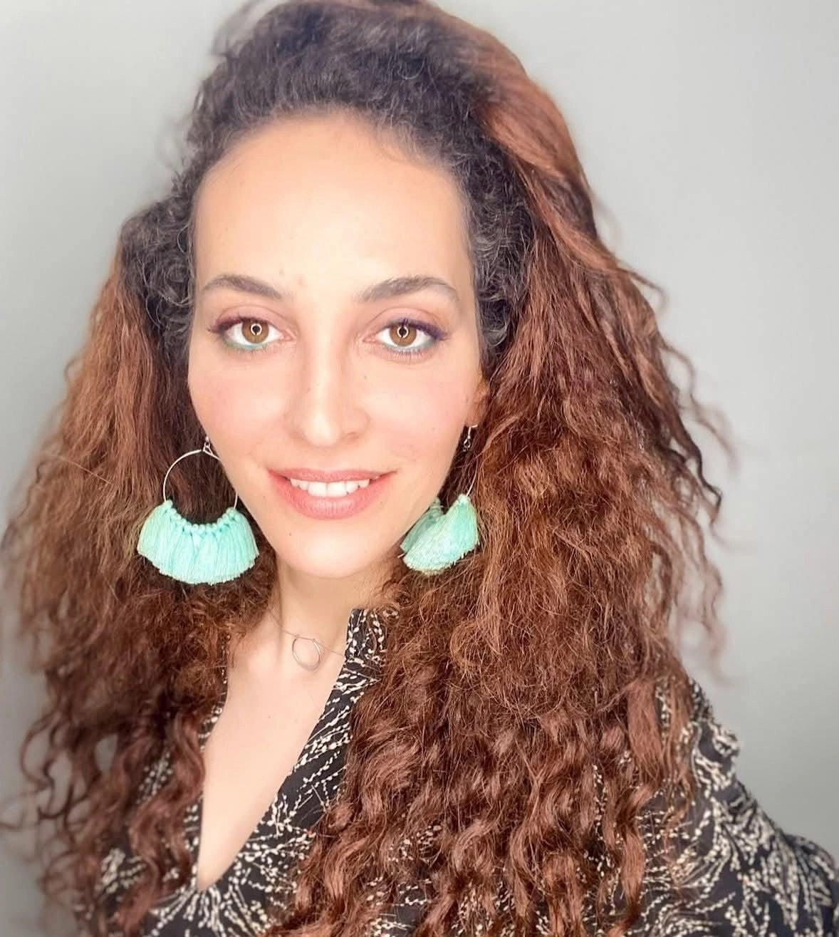 אישה עם שיער חום ארוך ומתולתל, עונדת עגילי ציצית גדולים בצבע תכלת ולובשת חולצה שחורה עם הדפס, מחייכת על רקע בהיר וחלק.