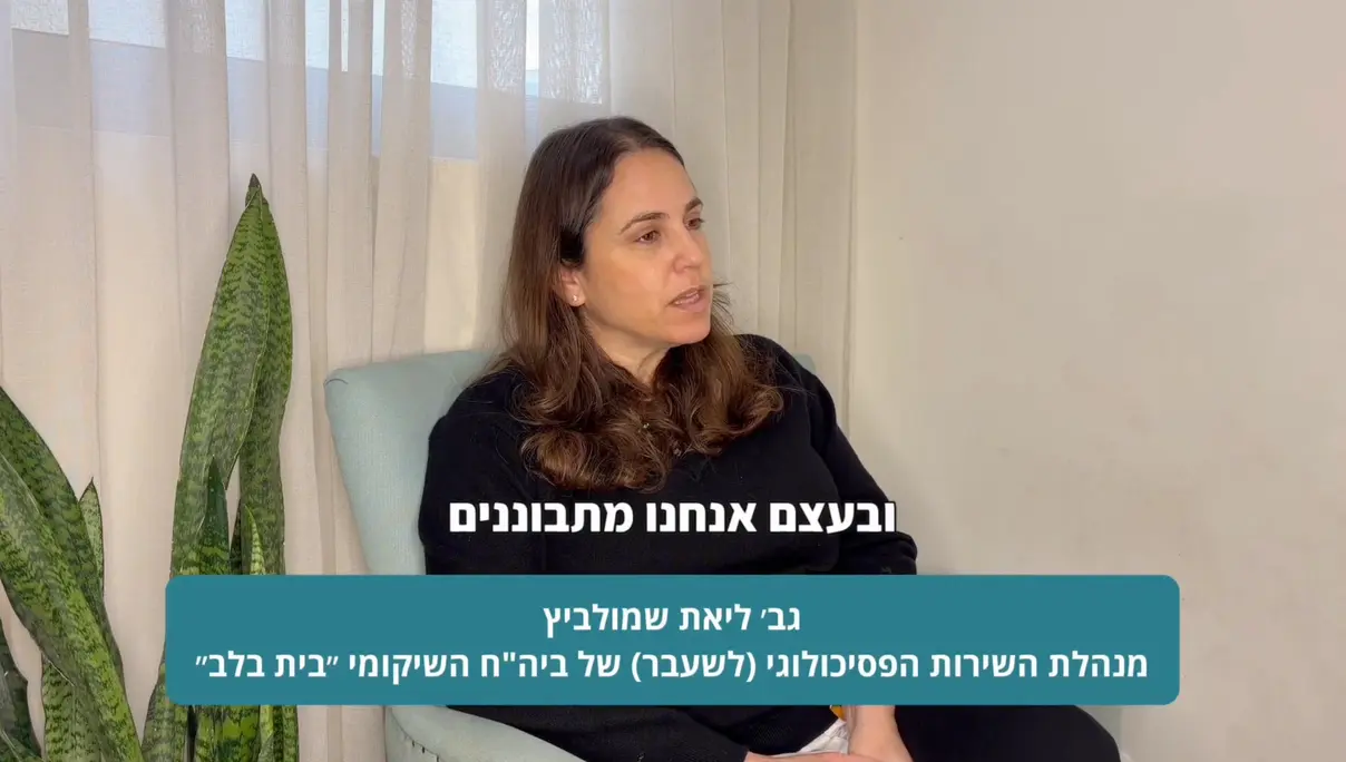 אישה עם שיער חום ארוך יושבת על כיסא ומדברת. לידה עומד עציץ. על המסך מוצג טקסט בעברית.